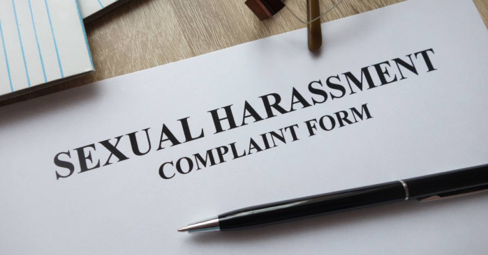 A sexual harassment compliant form lays on a wooden desk with a pen, a pair of glasses, and a notepad scattered around it.