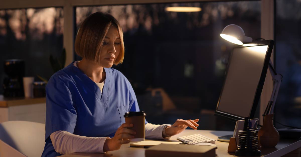 A woman wearing blue scrubs sits at a computer at night with a desk light on and holds a cup of coffee.