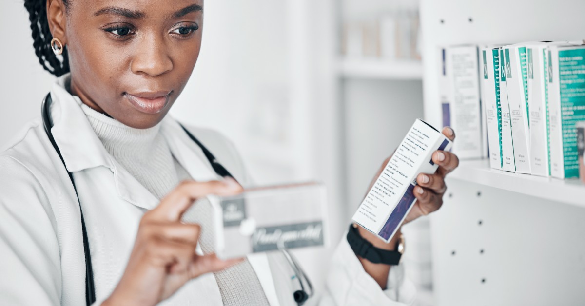 A woman wearing a doctor's coat and stethoscope around her neck holding up two boxes of pills and reading the labels.