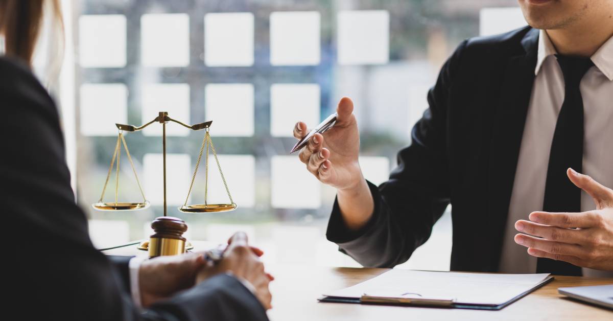  A focus on two men in black suits sitting at a desk with a clipboard. Scales and a gavel rest in the background.
