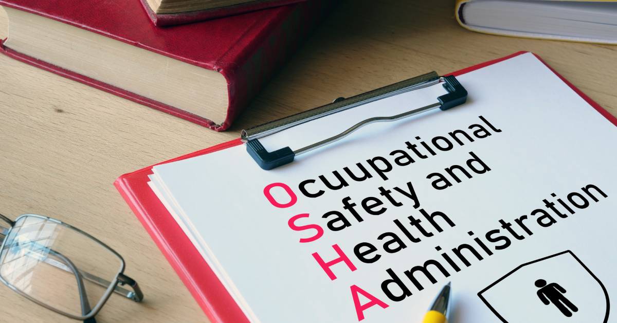 A red clipboard and books lie on a wooden desk. The clipboard reads "Occupational Safety and Health Administration."