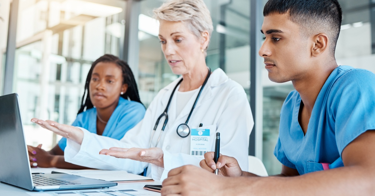 A doctor in a white coat sits in between two people wearing blue scrubs and taking notes in front of a computer.
