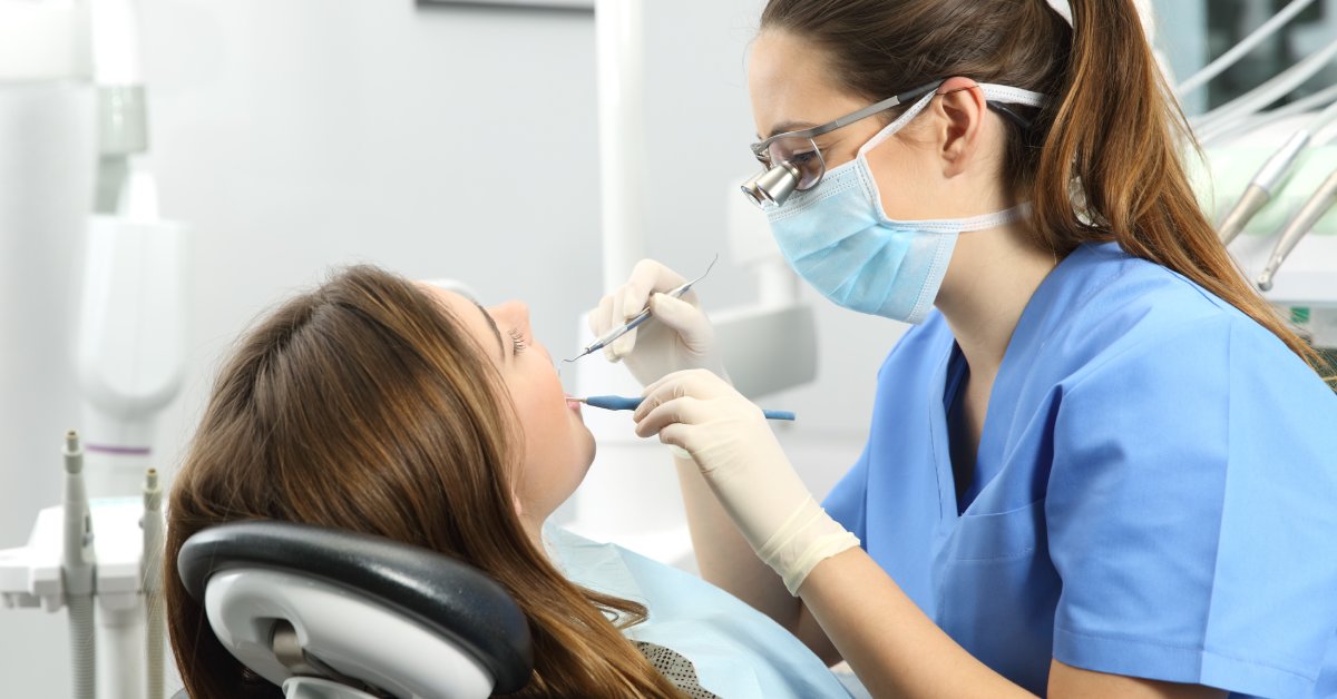 A female dental hygienist wearing a mask and blue scrubs examines a female patient's teeth in a dental office.