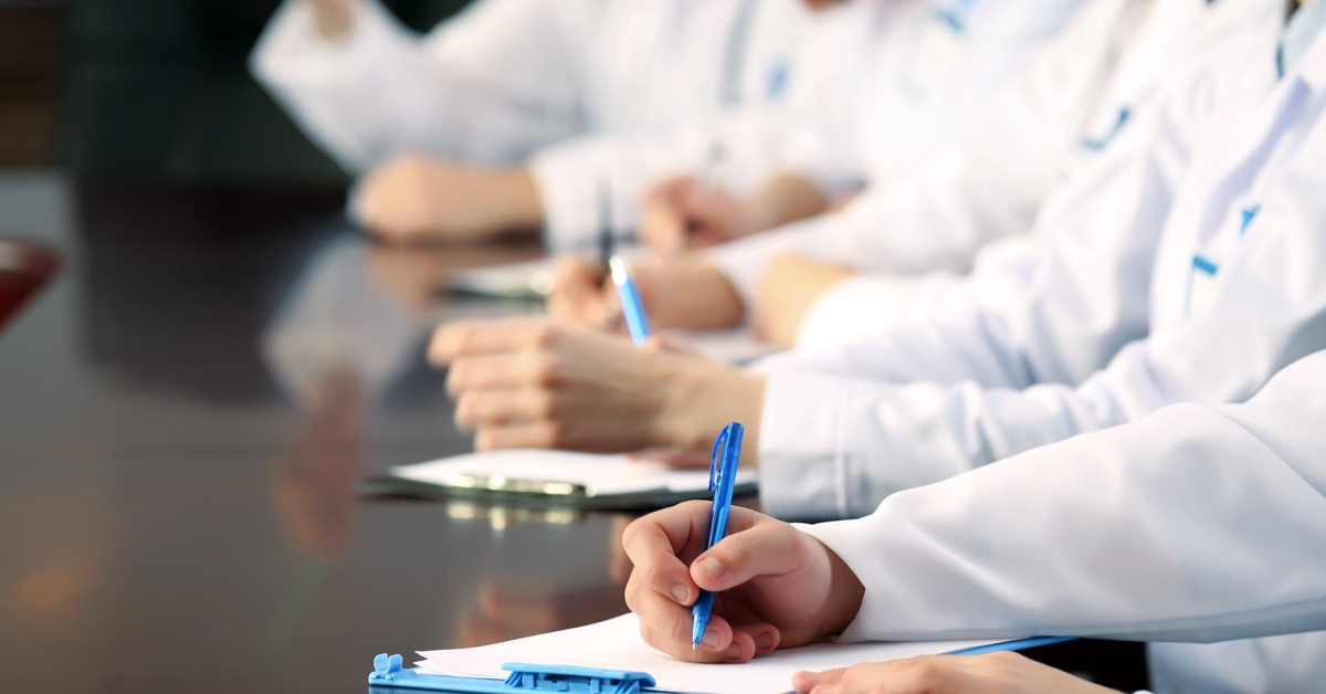 Several medical workers in white coats sit at a dark table with clipboards in front of them, taking notes.