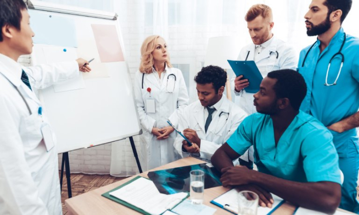 A group of people wearing scrubs and white coats gather around a table and a whiteboard with papers and notes.