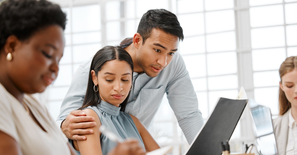 A male coworker puts his hand on an uncomfortable-looking female coworker's shoulder as he reads her report.