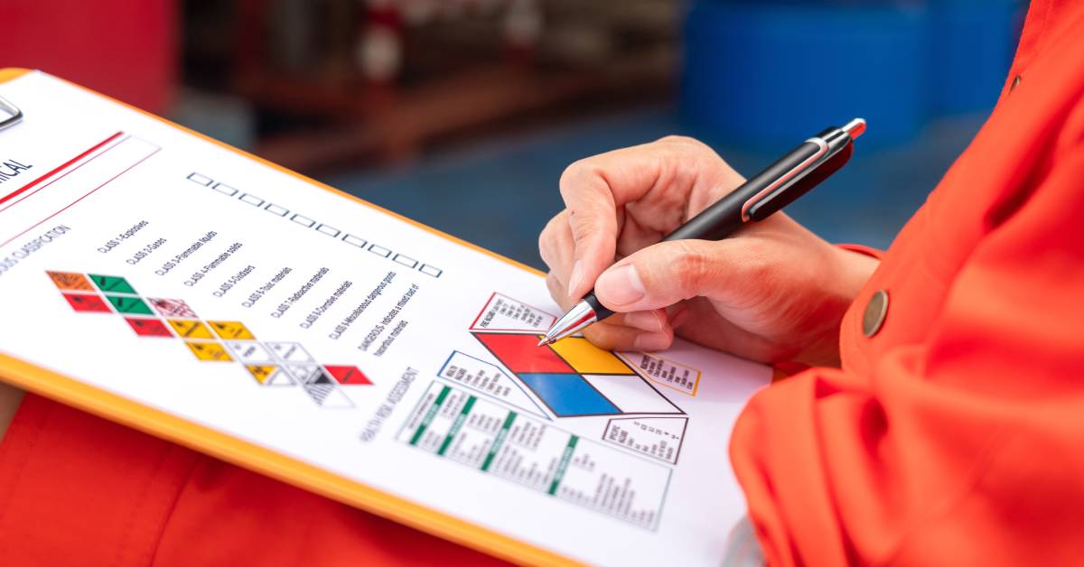 A person is holding a ballpoint pen and a clipboard that details different hazardous material classes.