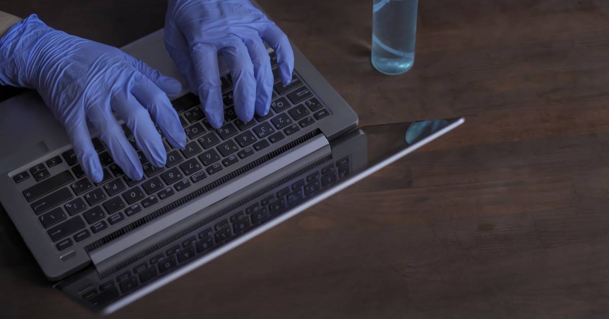 A pair of gloved hands typing on a laptop keyboard on a wooden desk with a bottled liquid sitting to the side.