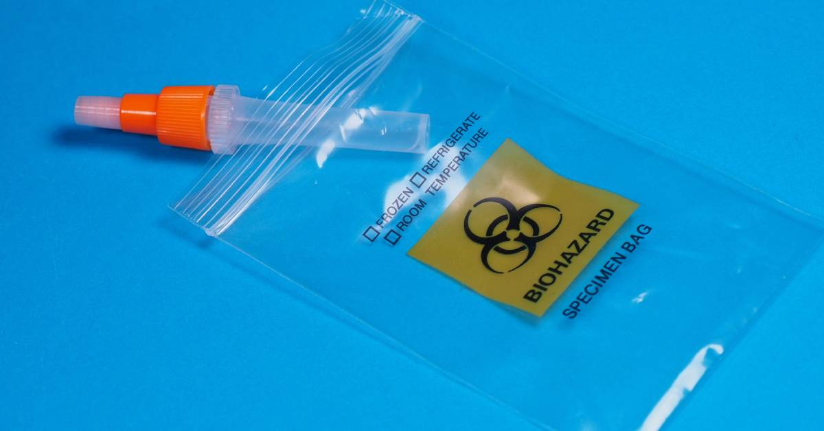 A small tube with an orange cap sits on a blue background, halfway in plastic bag with a biohazard symbol on it.