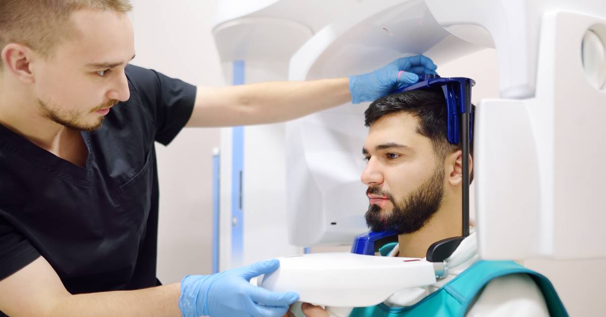 A dentist office staff operates an x-ray machine while a patient sits in its seat in a protective vest.